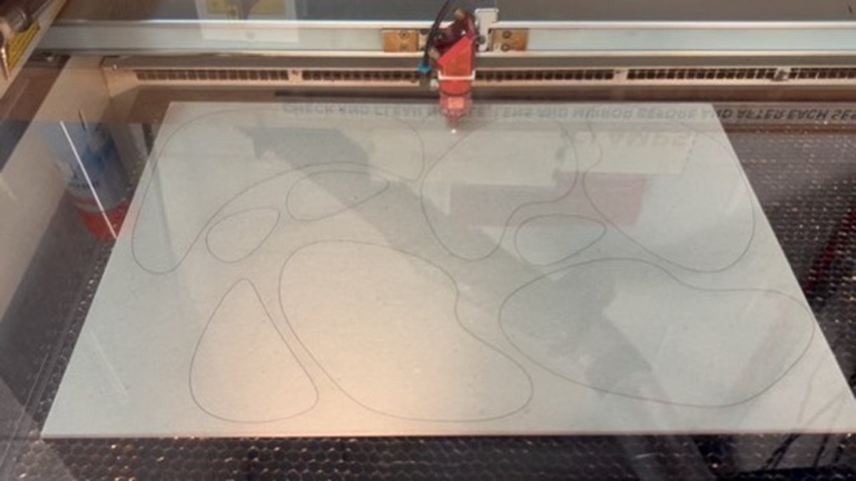 Overhead view of a laser cutter mid-cut, tracing layered contour lines into a sheet of material
