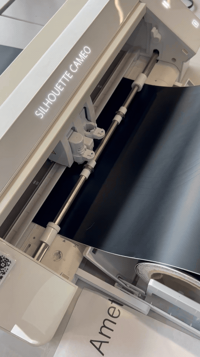 A Silhouette Cameo cutting machine running, feeding black vinyl material through the blade carriage