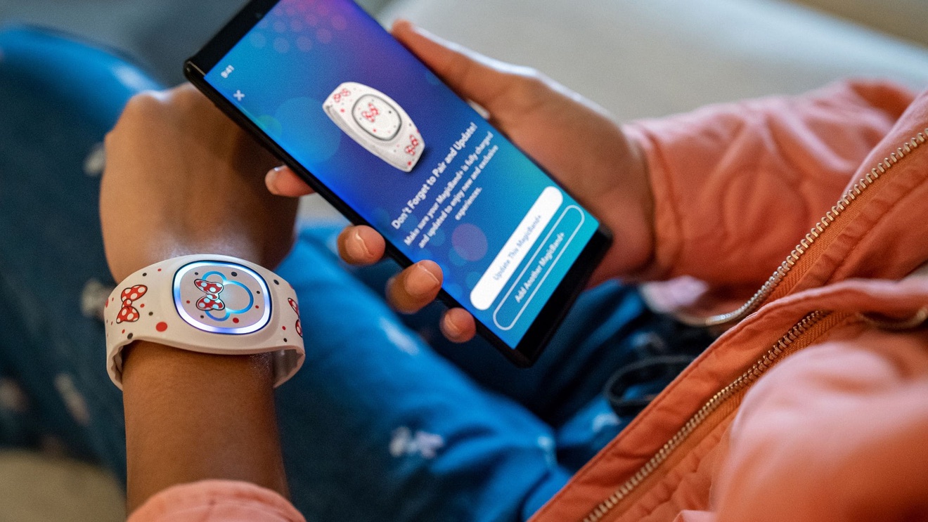 A guest holding a phone open to My Disney Experience beside a MagicBand on their wrist
